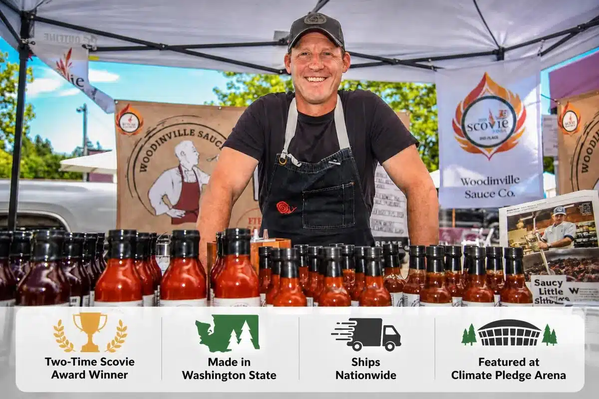 Smiling man in a black shirt and apron selling bottles of hot sauce at an outdoor market booth.
